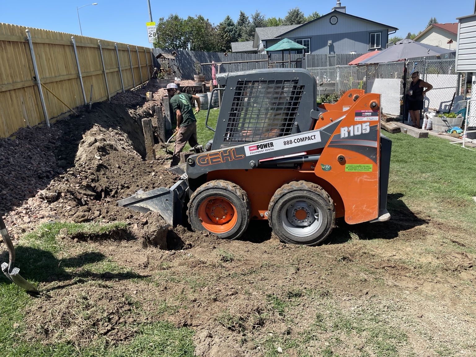 Gehl skid steer digging a rock trench along a backyard fence line